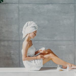 horizontal-shot-of-relaxed-young-european-woman-applies-moisturizing-body-cream-on-legs-wrapped-in_t20_A9zyR0